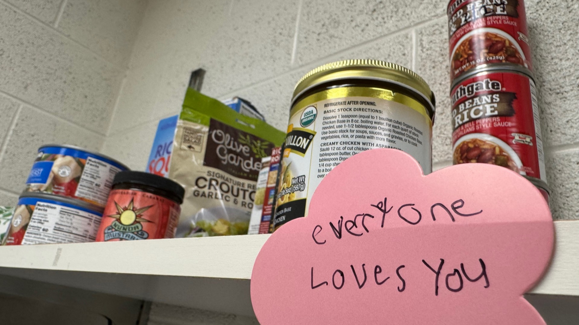 photo of a food pantry. A sticker adhered to a shelf, reads, in a child's handwriting, "everyone loves you".